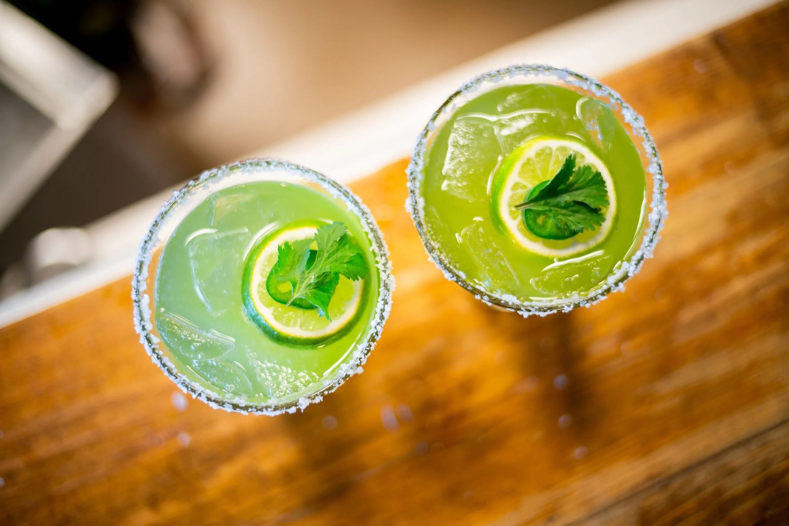 Two salt-rimmed green margaritas with lime slices and mint on a wooden table